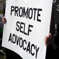 A protester holding a sign that reads PROMOTE SELF-ADVOCACY