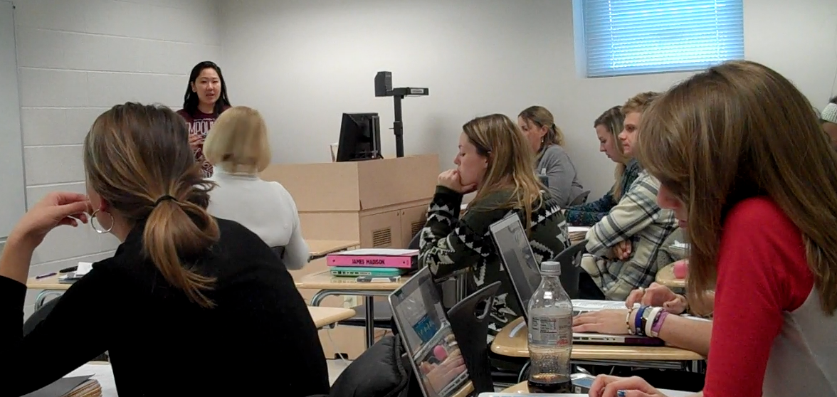 Image of a video screenshot depicting a classroom of college students at desks watching another student present