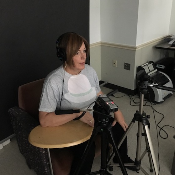 Joyce sits in a chair with headphones on, a Zoom mic recorder in front of her on a tripod, and another tripod in front of her with a webcam attached
