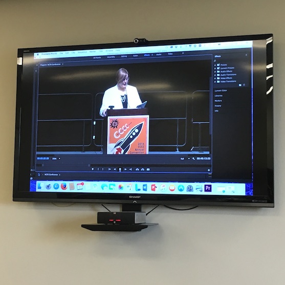 a wall-mounted television showing a draft of the video (with Joyce at a podium) in Premiere Pro