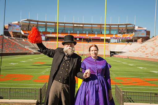 bearded white man in black suit and top hat and white woman in 19th century purple dress stand in front of a football field