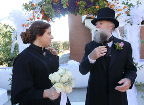 a woman with dark hair, wearing a black dress and holding a bouquet, looks at a man in a black suit and top hat with a large beard