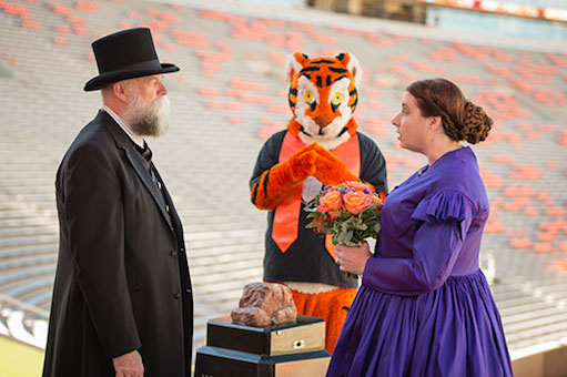 man and woman dressed in old-style clothes face each other; she holds boquet as Clemson Tiger mascot stands between them like a priest marrying them