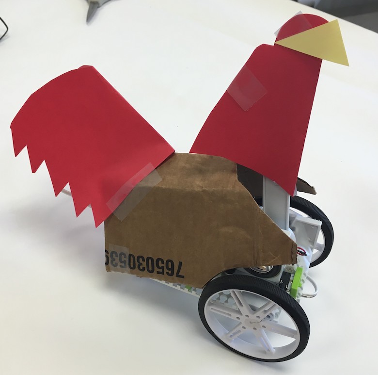 a littleBits construction of a chicken: over a littleBits invention (with two visible wheels) are a cardboard body and red and yellow construction paper for a tail and head