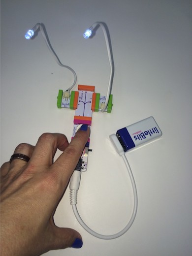 a littleBits construction with two lights at the end of wires sticking up like alien eyes; a person's hand presses a button attached to the littleBits invention