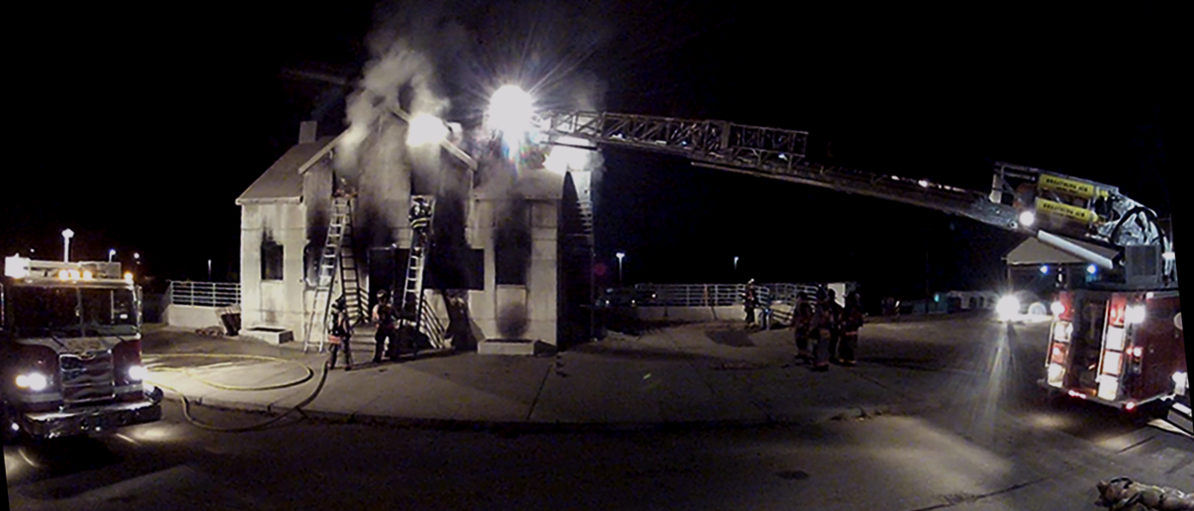 Members of an engine and a ladder company practice coordinated fire attack at a live-fire training facility. A hoseline has been stretched from a fire engine through the front door of the two-story concrete structure. Smoke issues out from the second floor, as members of the truck company climb ground ladders. The tower ladder has been extended to the corner of the structure.