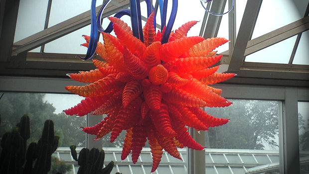 Photo of a glass sculpture descending from a glass ceiling. The sculpture is shaped like a red plant with a blue stem.