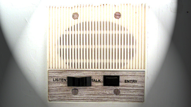 A white intercom box, mounted on a wall. The top of the box is a speaker, and the control panel sits on the bottom. There are three buttons: listen, talk, and entry.