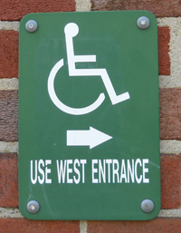 A disability sign with the iconic wheelchair image, nailed to the wall of a brick building. The text reads USE WEST ENTRANCE and has an arrow pointing to the right.