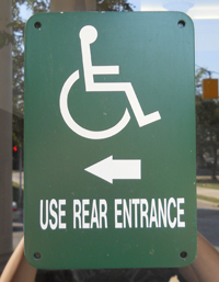 A disability sign with the iconic wheelchair image, hung on a glass door. The text reads USE REAR ENTRANCE and has an arrow pointing to the left.
