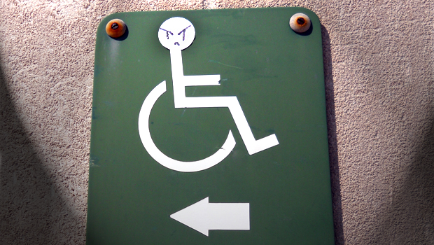 A building sign with the iconic wheelchair figure. Someone has drawn an angry face on the wheelchair user.