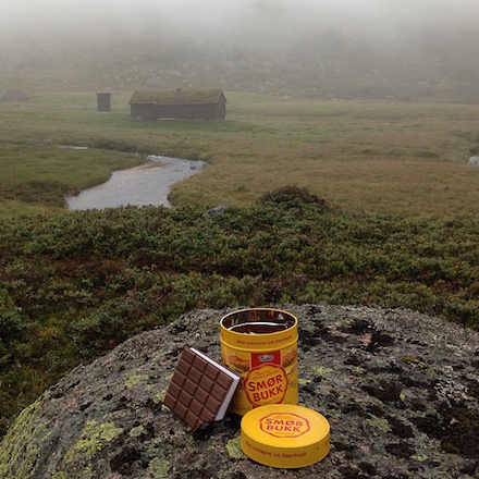 An unusual cache container—a small yellow tin with a plastic representation of an ice cream sandwich—is positioned on a rock in the foreground of the photograph, with a foggy green marsh and brown farm buiding in the background.