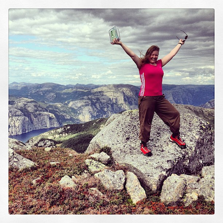 A woman triumphantly holds a geogache container aboive her head, standing atop a rocky outcrop with the dramatic cliffs of a Scandinavian fjord in the background.