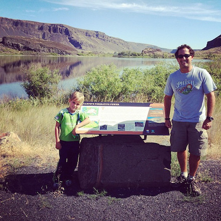 A man, to the right of the frame, stands next to a natural landmark sign, with a large lake and desert cliffs in the background. A young boy with blond hair and a bright green Seattle Sounders jersey stands to the left of frame, on the other side of the sign.