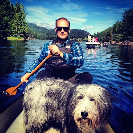 A man paddles a canoe on a gorgeous lake as his shaggy dog stands in front of him.