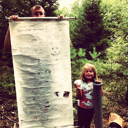 An unusual cache log: a man, to the left of frame, holds a giant scroll with both hands at nose height, the log itself covering his whole body and rolling to the ground. A young girl stands next to him, to the right of frame, holding the large tube that is the cache container.