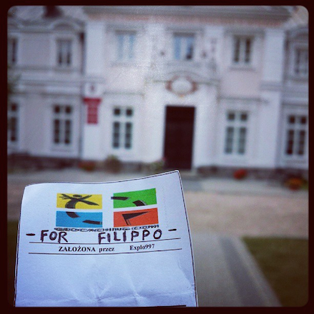 The photographer holds a clean cache log (one that has not been signed); this is significant because this cache has remained unfound for 4 years and he is the first to find it. A large, ornate building is in the background.