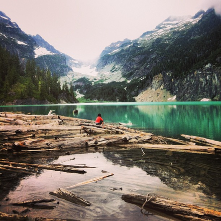 A woman in a red sweatshirt sits at the edge of a small emerald green lake in the midst of snow-capped peaks.