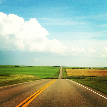 A lonely country road stretches into the distance, framed by high clouds, blue sky, and green and yellow farm fields.