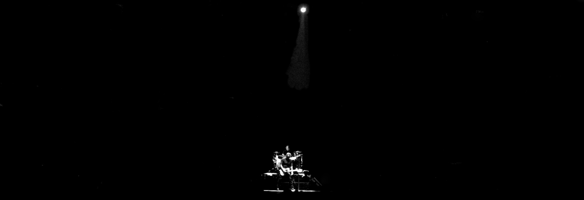 black and white photograph taken from the distance of Springsteen on a darkened stage with one light shining on him