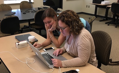 Erica sits at a table in a classroom working on her MacBook Pro; Sarah leans over her right shoulder, smiling at the laptop screen