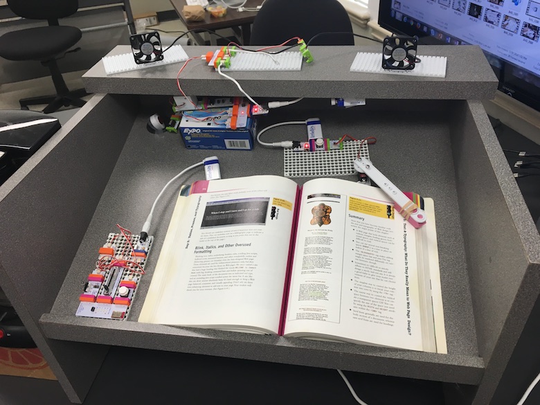 sitting on top of a podium are a bunch of littleBits (fans, wires, batteries, small lights, and more) and an open textbook with a mechanical arm resting on the right page