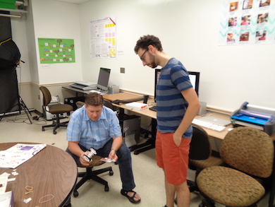 Rick is sitting, holding the remote control car and studying it; Jack stands beside Rick looking down at the invention