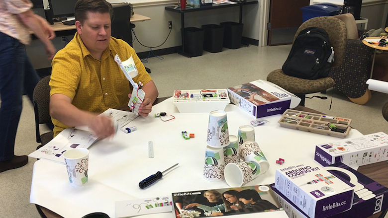 a man on the left holds the littleBits throwing arm invention, launching a paper ball toward a tower of plastic cups, with some of the cups already knocked over