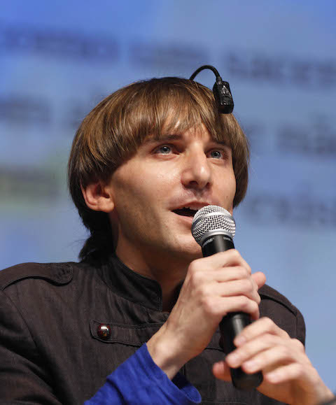 Harbisson, a white man with medium-length hair, holds a microphone and looks up at his antenna attached to his skull