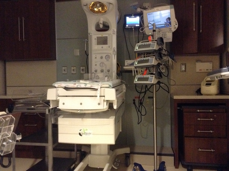 A bed space in a pod is set up for an admission to the NICU. The bed is surrounded by medical equipment that reaches over 7 feet tall.