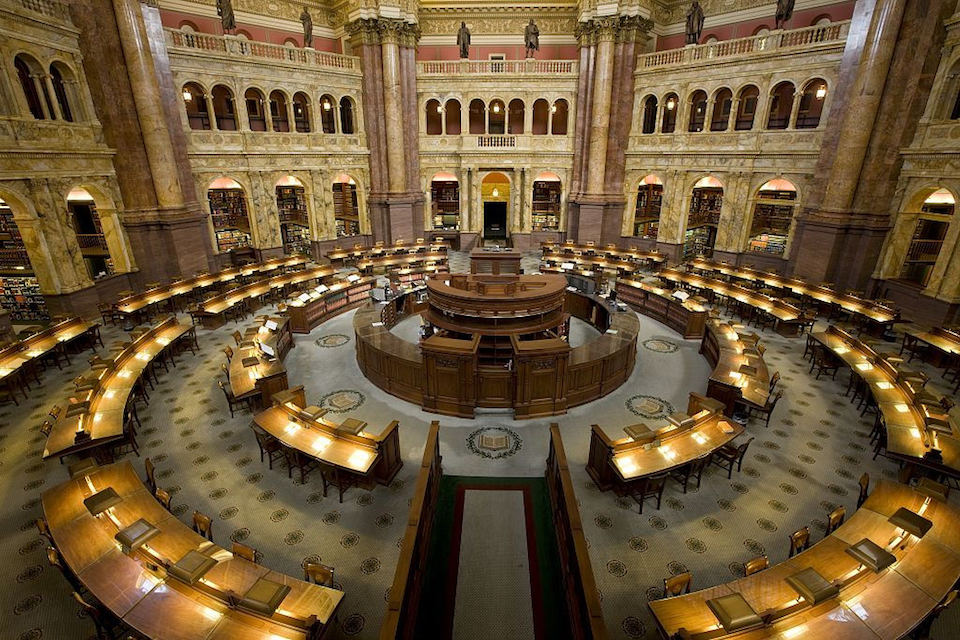 The Library of Congress Reading Room is shown without any people. The working spaces are desks formed in three concentric circles that emanate from the reference librarians' circular desk in the center of the over two-story room. The floor is carpeted, one assumes, to disrupt sound echoes.