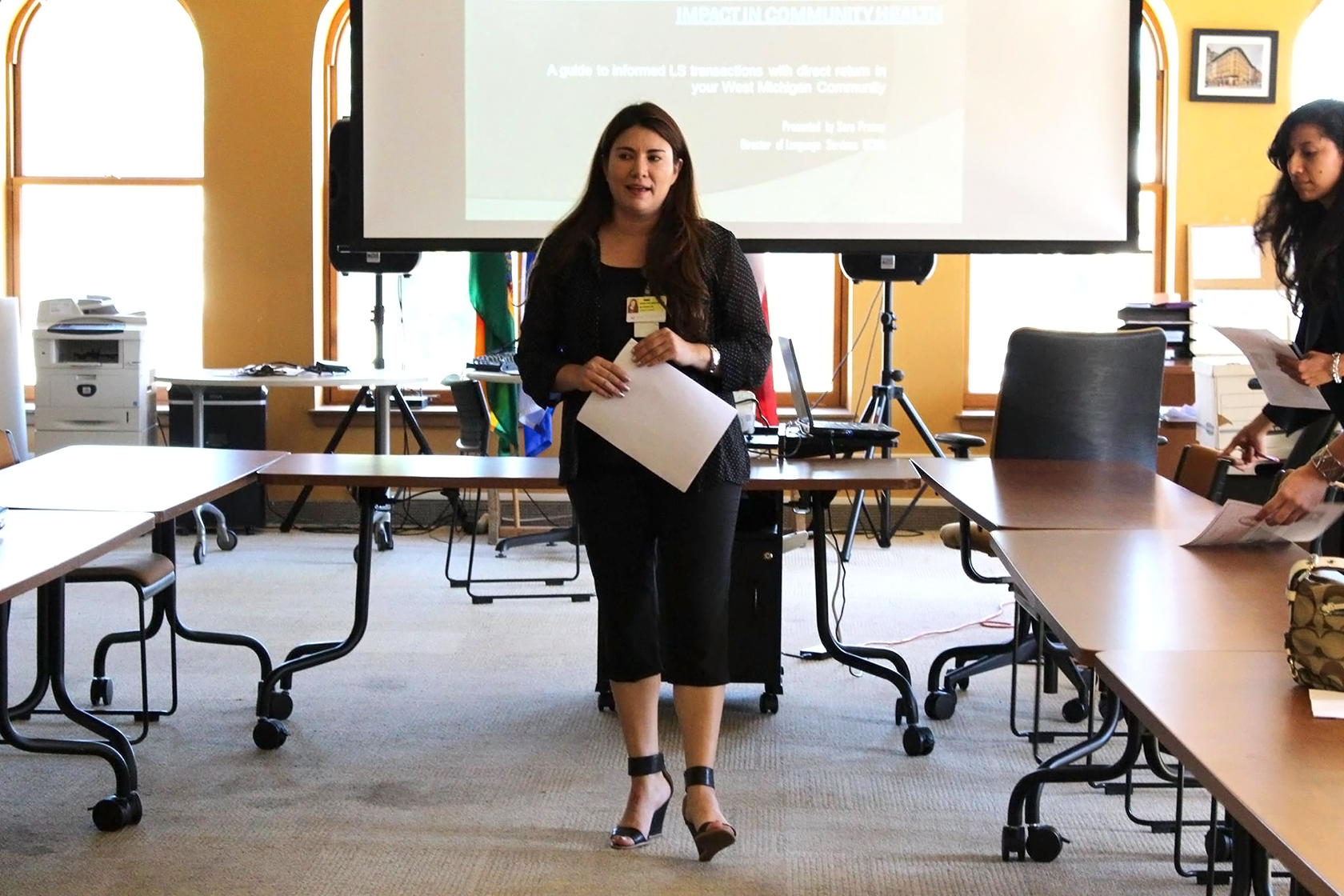 Former Language Services Director Sara Proano stands in front of a projector and in the center of several rectangular tables for workshop participants forming a square around her while holding a document.