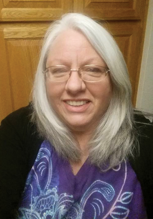 A photo shows a woman with light skin and gray hair who stands smiling in front of a door.