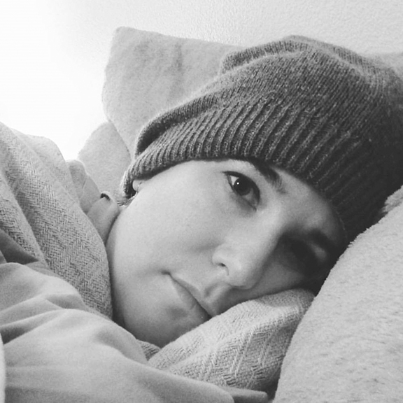 A black-and-white image of a woman lying in bed wearing a toque and facing the camera.