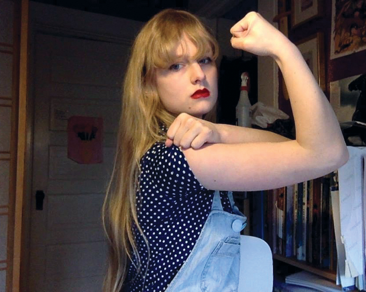A photo shows a woman in overalls, a polka-dot shirt, and red lipstick. She is flexing her right bicep.