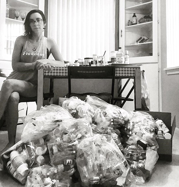 Black-and-white image of a woman seated at a table with a heap of garbage bags in front of her containing discarded pill bottles.