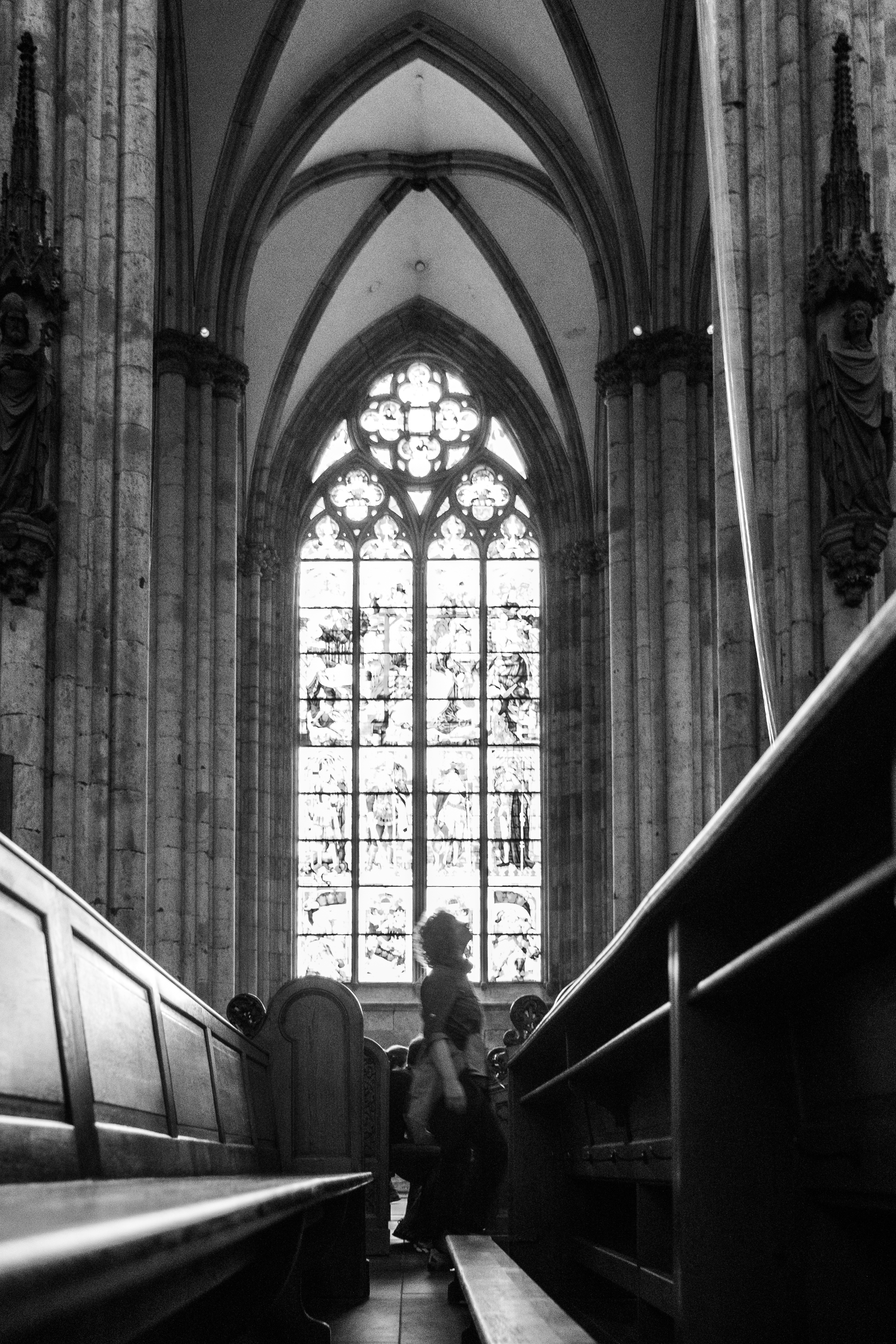 A solitary figure stands in a church with flying buttresses under a stained glass window