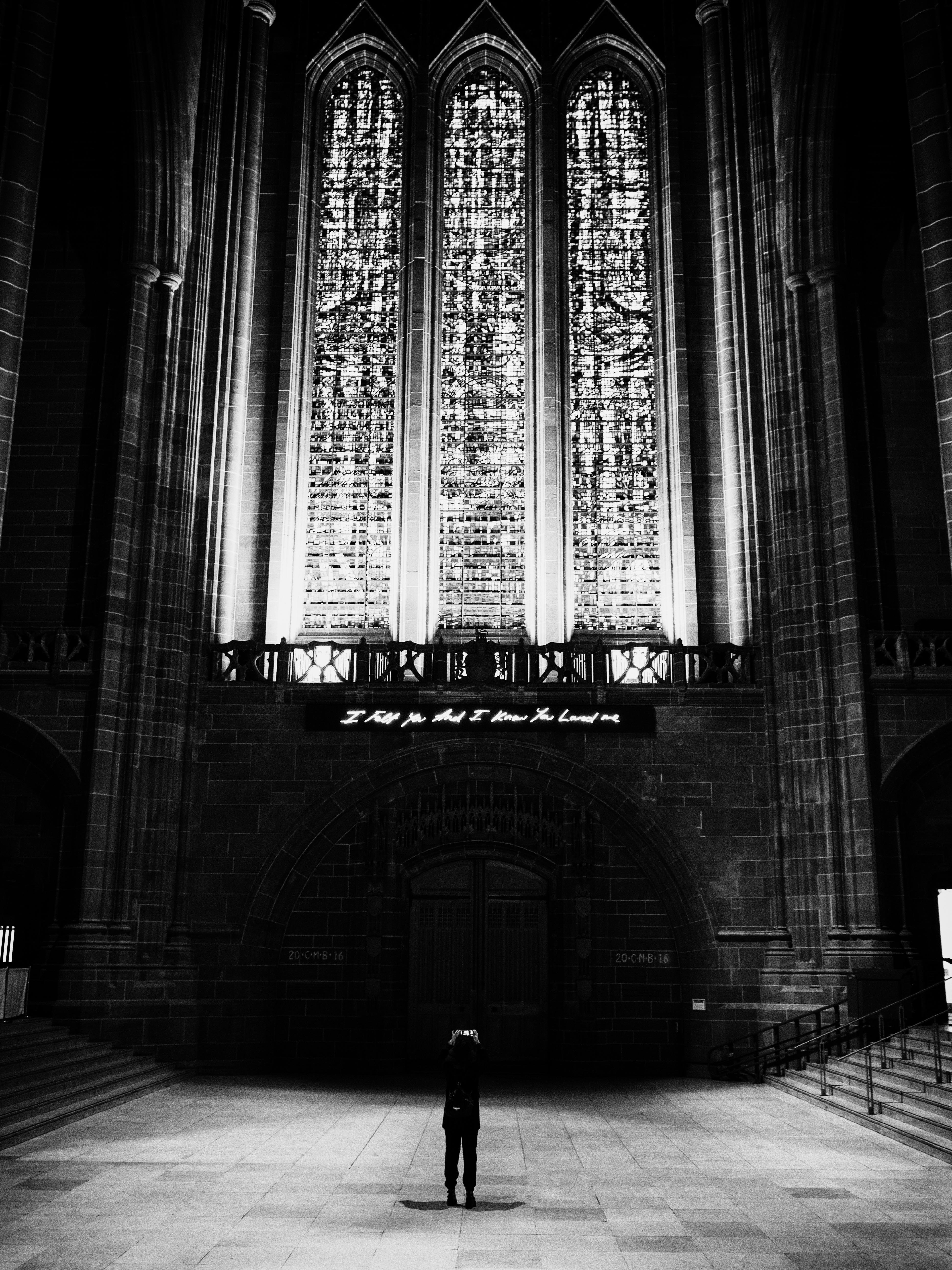 A solitary figure stands in a church in front of three towering stained glass windows