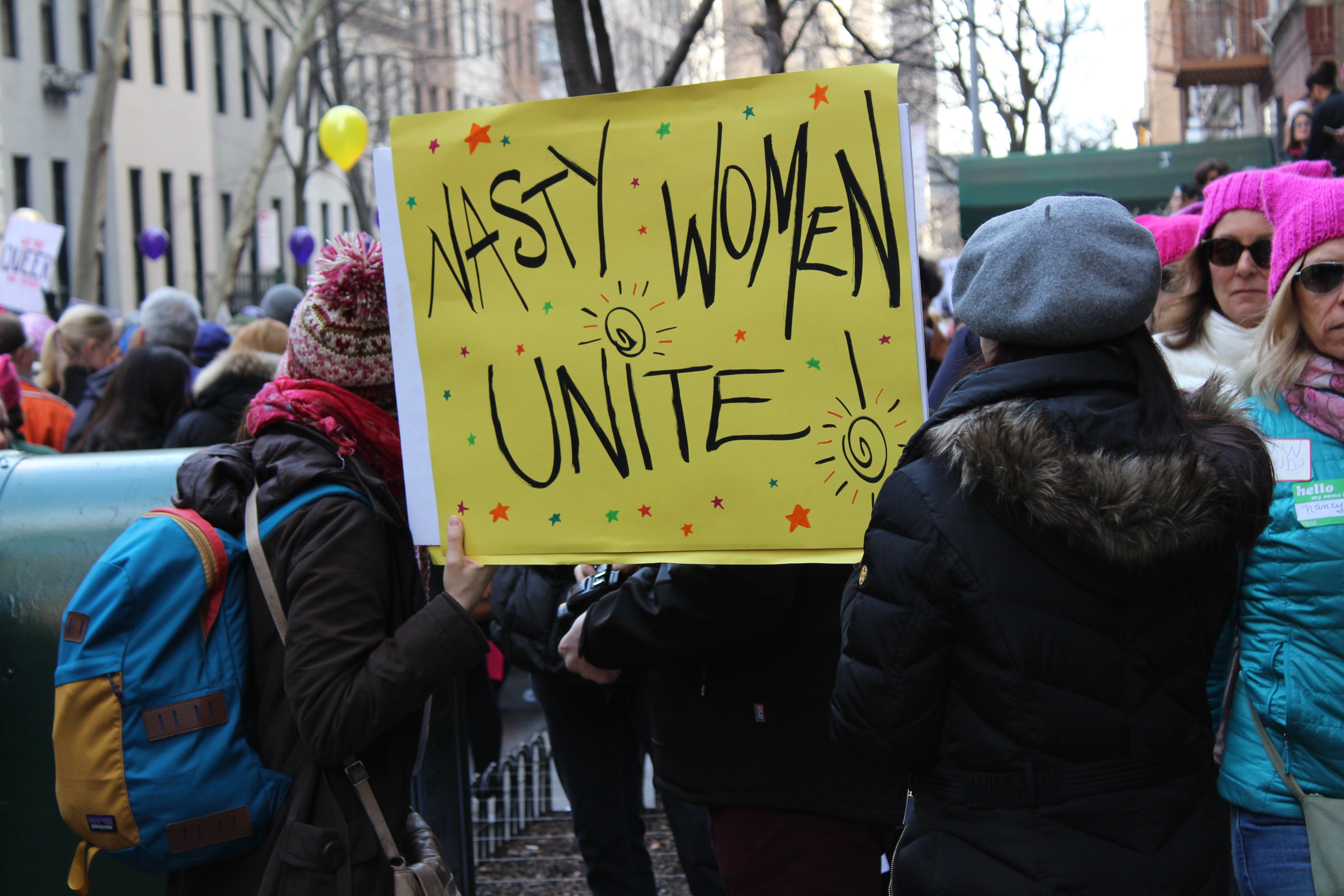 Protesters holding a sign that says Nasty Women Unite! There are women wearing pink knitted pussy hats in the background