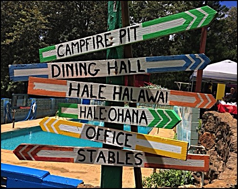 Green pole with six different colored arrows stacked vertically pointing in different directions indicating directions for locations at Camp Palehua, Hawai'i including Campfire Pit, Dining Hall, Hale Halawai, Hale: Ohana, Office, and Stables.