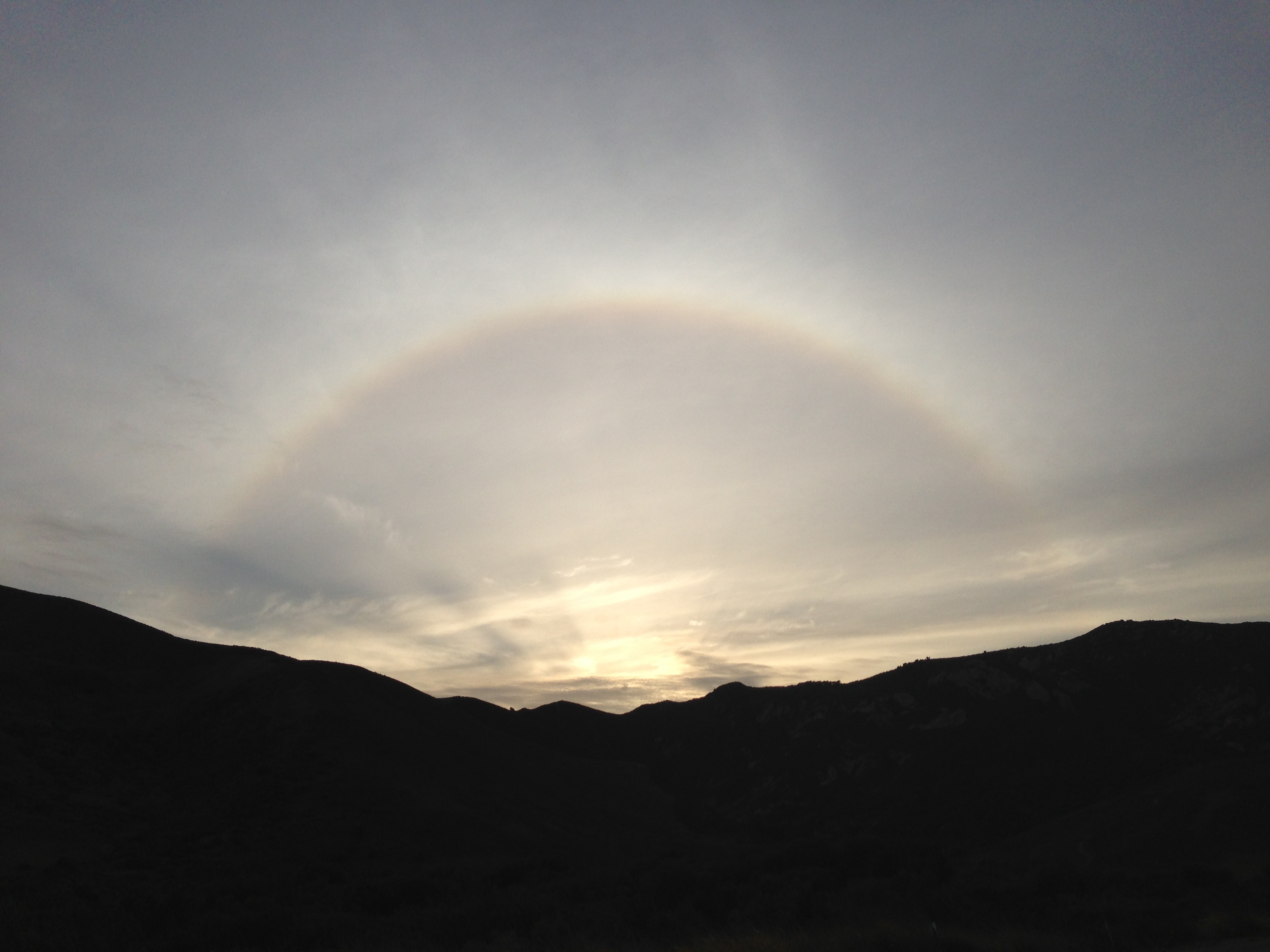 A faint rainbow, or sundog, spanning a dark silhouette of mountains beneath clouds in an evening sky.