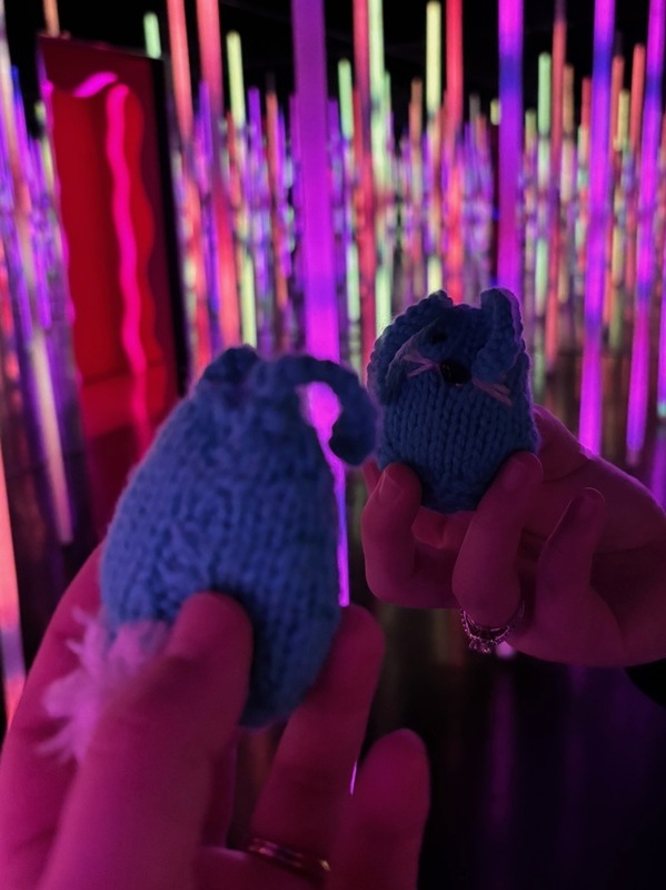 A small blue knit bunny looks at herself in the mirror. Purple, pink, and green tubes glow in the background.