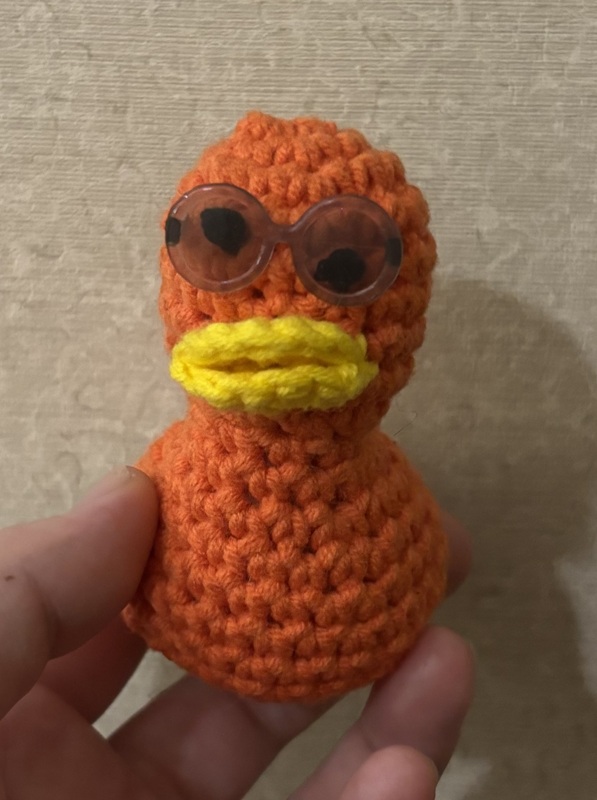 A hand holds an orange crocheted duck wearing grey sunglasses. One eye is higher on its face than the other.