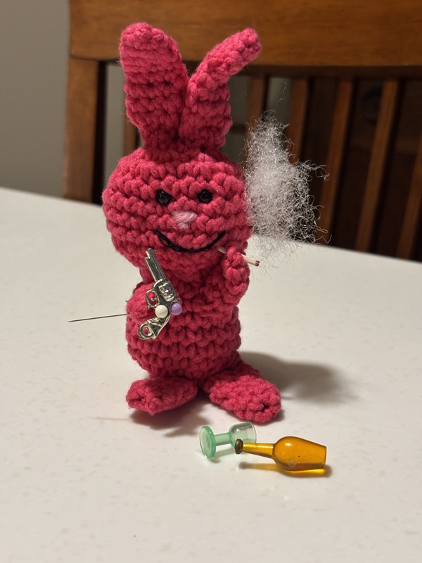 Evil Bunny holds a tiny gun in his right hand and is bringing a cigarette up to his mouth with his left. A plume of 'smoke' made from fiberfill stuffing rises from the cigarette. A glass a bottle are toppled over at the bunny's feet.