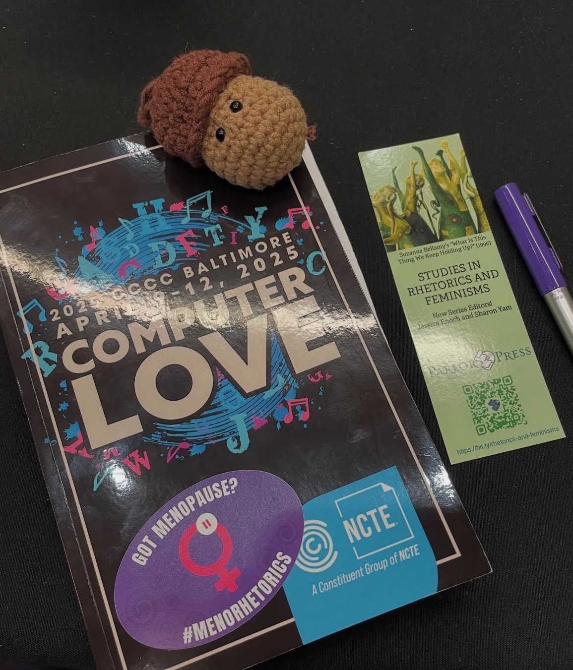A brown crochet acorn, a green bookmark for Studies in Rhetorics and Feminisms, and purple pen, and a CCCC2025 program book with a Got Menopause? sticker all arranged on a black background.