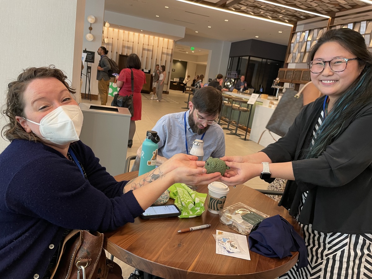 Hannah extends a the first conference creature, a forest green four-legged creature with ears, across a table in a conference hotel lobby, to Cagle.