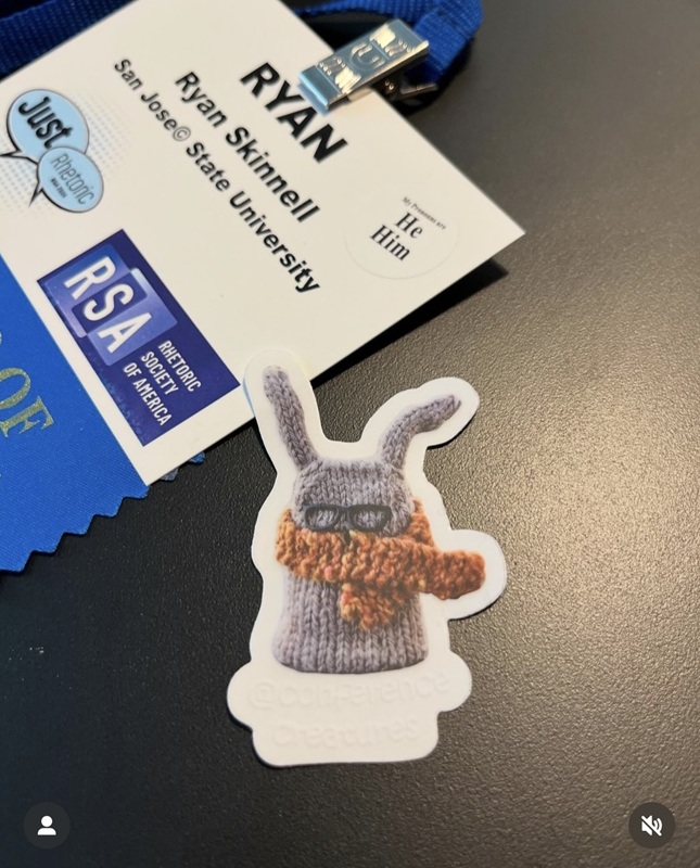 An un-stuck Conference.Creatures sticker with Ryan Skinnell's RSA name badge behind it. Posted to instagram by @rhetoric_sticklies.