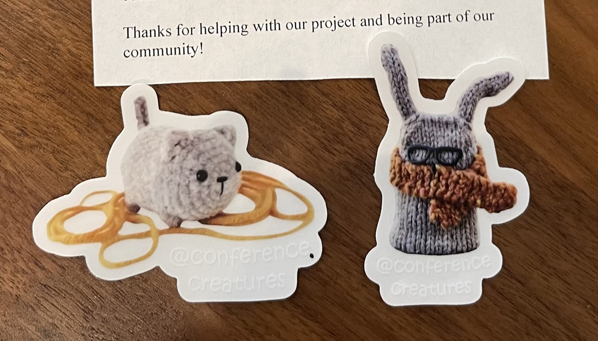 Two stickers, one of a crochet cat standing on yellow yarn and the other with a grey bunny wearing a brown scarf and black glasses, both with the handle @conferencecreatures is printed in white beneath the bunny.
