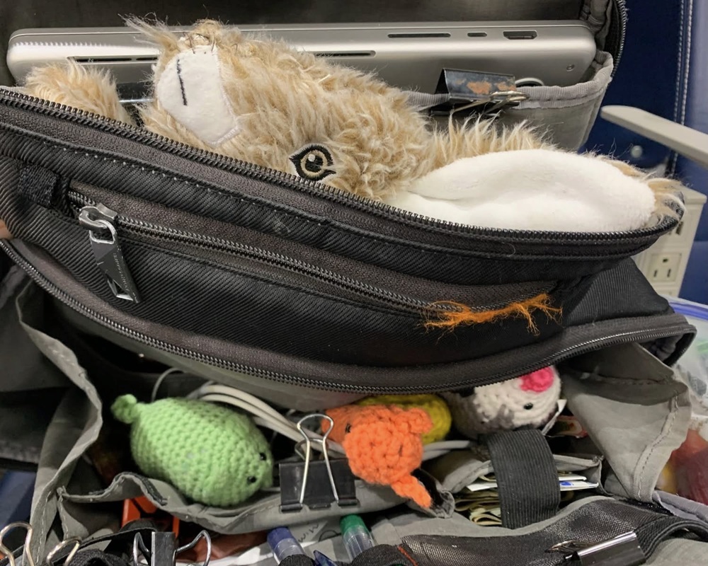A brown fox cat toy, a light green crochet cat, a crochet possum, and a crochet snail poke out of a gray travel bag on an airplane.