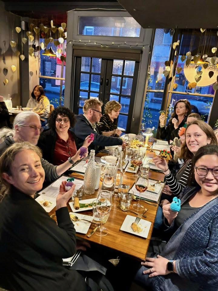 Nine writing studies scholars sit around a dinner table at a Thai restaurant and hold up various crochet creatures for the camera.